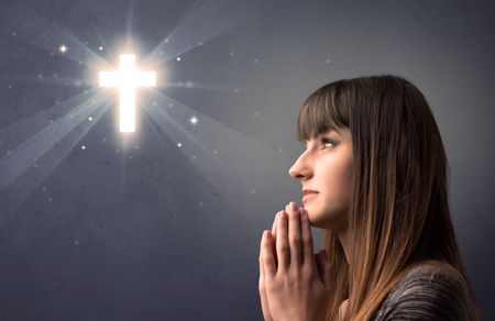 Young woman praying on a grey background with a shiny cross above her