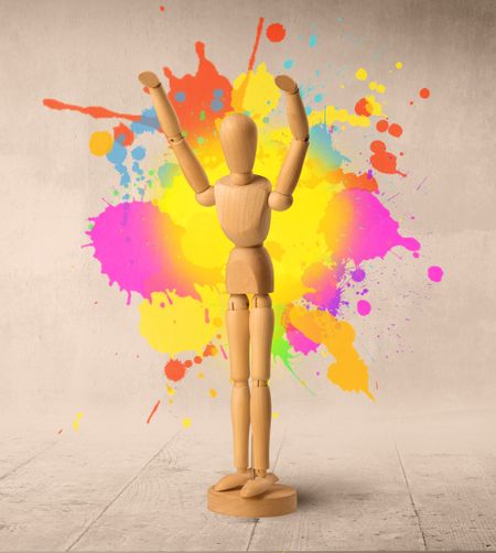 Wooden mannequin posed in front of a greyish background with colorful splashes behind it