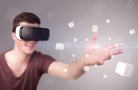 Young impressed man wearing virtual reality goggles with grey cubes around him 