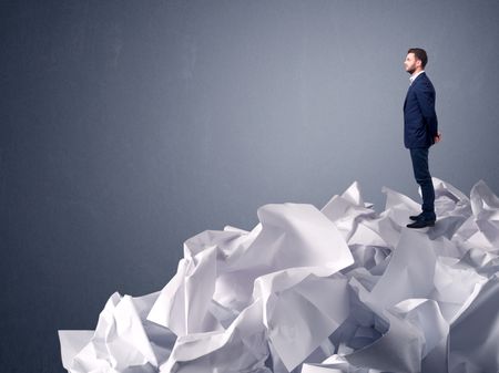 Thoughtful young businessman standing on a pile of crumpled paper with a light grey background