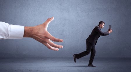 Young miniature businessman running from a big hand with blueish grey background