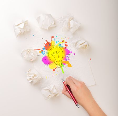 Female hand next to a few crumpled paper balls drawing a colorful lightbulb