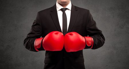 A well dressed sales person standing with red boxing gloves on his hand in front of urban grey wall background concept.