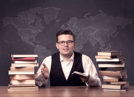 A young ambitious geography teacher in glasses sitting at classroom desk with pile of books in front of world map drawing on blackboard, back to school concept.
