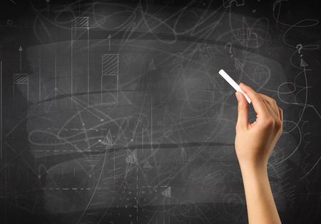 Female hand holding white chalk in front of a blackboard with scribbles and plans drawn on it 