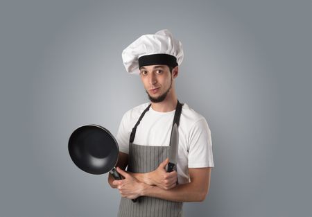 Young bearded cook portrait with kitchen tools and empty wallpaper