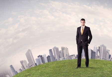 A successful male good looking business man standing in small green grass in front of city landscape with skyscrapers concept.