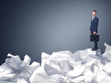 Thoughtful young businessman standing on a pile of crumpled paper with a blueish grey background