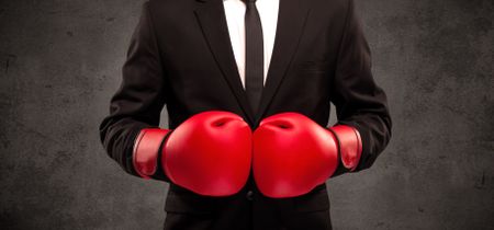 well-dressed-sales-person-standing-red-1059749528 A well dressed sales person standing with red boxing gloves on his hand in front of urban grey wall background concept.