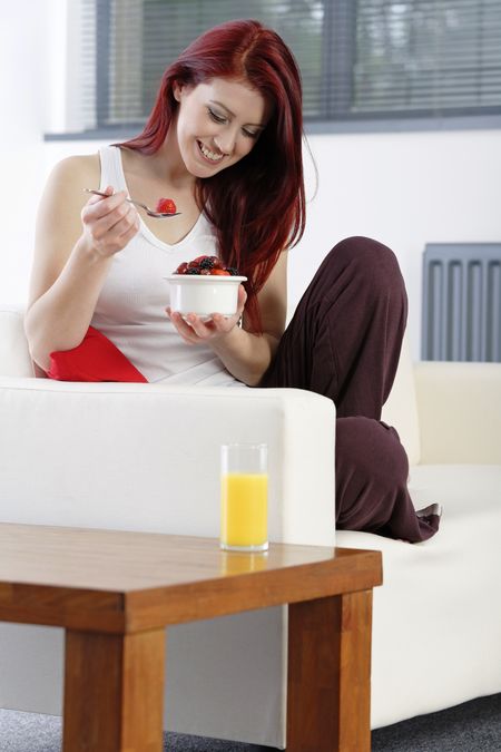 Happy young woman sat on her sofa at home enjoying a bowl of fresh fruit