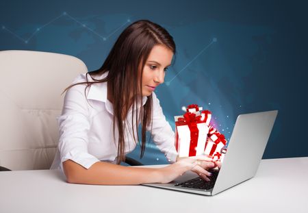 Pretty young lady sitting at desk and typing on laptop with present boxes icons