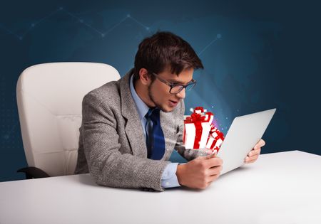 Angry young man sitting at desk and typing on laptop with present boxes icons