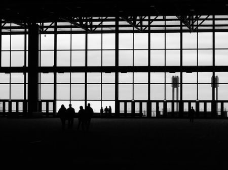 Eight unidentifiable adults in silhouette by closed doors and large windows of dark hall in convention center, in black and white