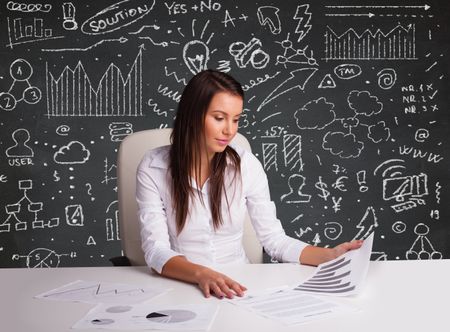Pretty young businesswoman sitting at desk with business scheme and icons