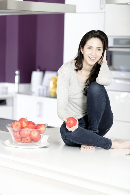 Beautiful young woman eating an apple in her white kitchen relaxing
