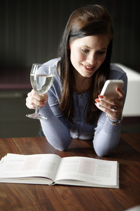 Attractive young woman enjoying a glass of wine in her kitchen while reading a text on the phone.
