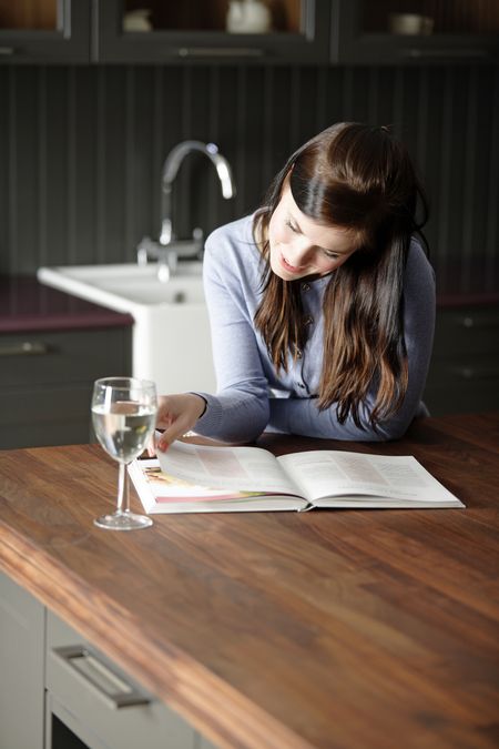 Attractive young woman reading a recipe from a cookery book in her kitchen with a glass of wine.