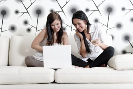 Two attractive young friends catching up at home on the sofa with a glass of wine using a laptop computer.