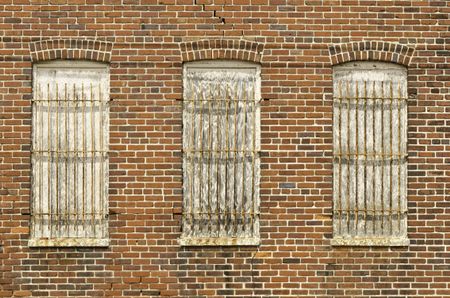 Industrial vintage: Three boarded windows, each with a rusty grate, on red brick exterior wall with cracks