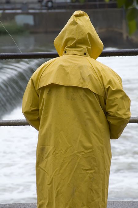Fisherman in yellow raincoat watches white river water over dam