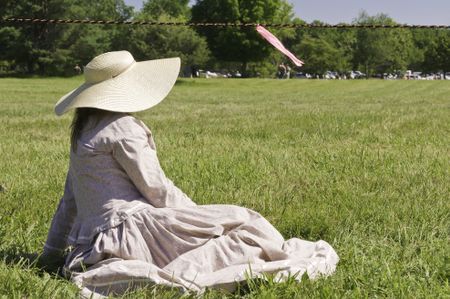 Unidentified woman actor in sun hat and period dress sits waiting by roped-off battlefield during reenactment of battle in the American Civil War (1861-1865)