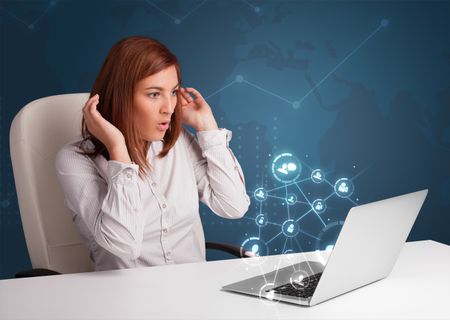 Beautiful young lady sitting at desk and typing on laptop with social network icons comming out