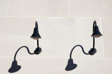 Two black light fixtures, each with a shadow, on exterior concrete wall in bright sunlight