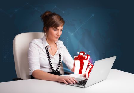 Pretty young lady sitting at desk and typing on laptop with present boxes icons