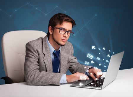 Attractive young man sitting at dest and typing on laptop with message icons comming out