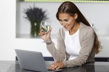 Young woman in her kitchen using a laptop while enjoying a glass of wine.