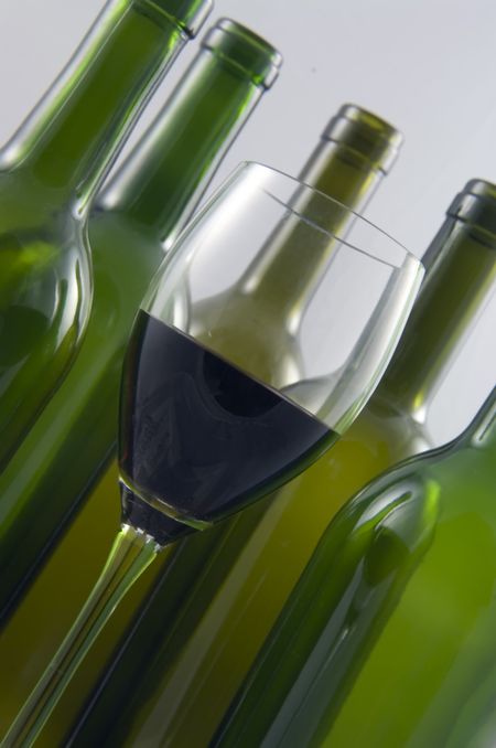 Glass of red wine with five green wine bottles in background, all tilted