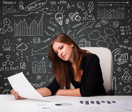 Pretty young businesswoman sitting at desk with business scheme and icons