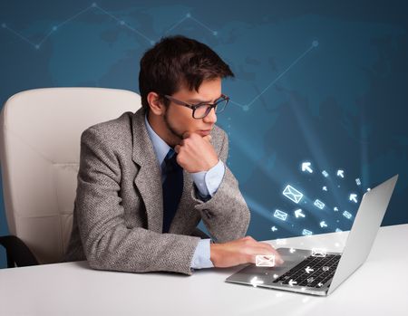 Attractive young man sitting at dest and typing on laptop with message icons comming out