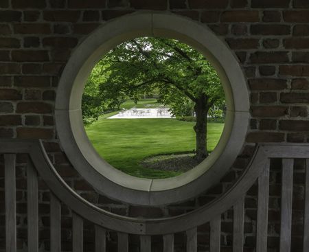 Outside world at a glance: Four-part porthole in a walled garden with a view of spacious green lawn, trees, and patio across the way