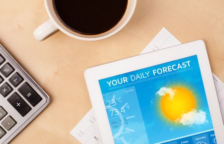 Workplace with tablet pc showing weather forecast and a cup of coffee on a wooden work table close-up