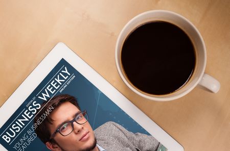 Workplace with tablet pc showing magazine cover and a cup of coffee on a wooden work table close-up