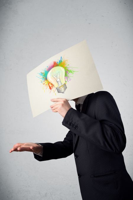 Businessman holding a cardboard with coloured paint splashes and lightbulb in front of his head