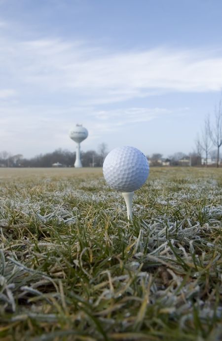 Elevated balls in perspective: golf ball teed up on frosty field with water tower on horizon