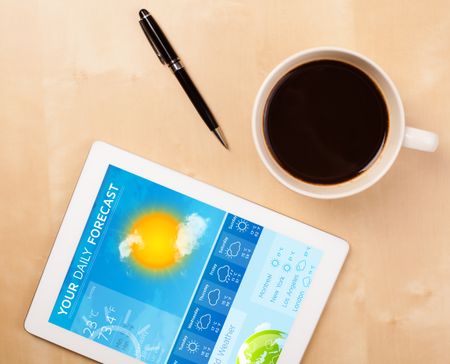 Workplace with tablet pc showing weather forecast and a cup of coffee on a wooden work table close-up