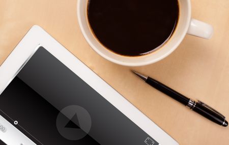 Workplace with tablet pc showing media player and a cup of coffee on a wooden work table close-up