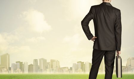 Businessman from the back in front of a city view with clouds and grass