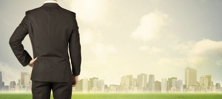 Businessman from the back in front of a city view with clouds and grass