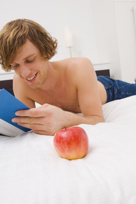 Guy reading a book on bed with an apple in the foreground.