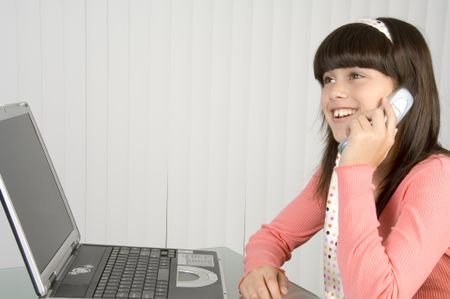 Pretty brunette schoolgirl in pink talking on cell phone, with bright smile, sitting by notebook computer and blinds