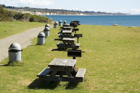 Row of picnic tables, grills, and garbage cans in public park along coastline