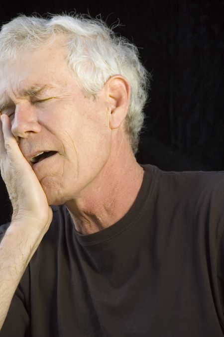 Mature white man with gray hair, eyes closed, head in right hand, mouth open, with expression of pain or regret or dismay