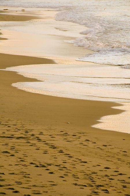 Ocean waves reflect light of setting sun on sandy beach with footprints