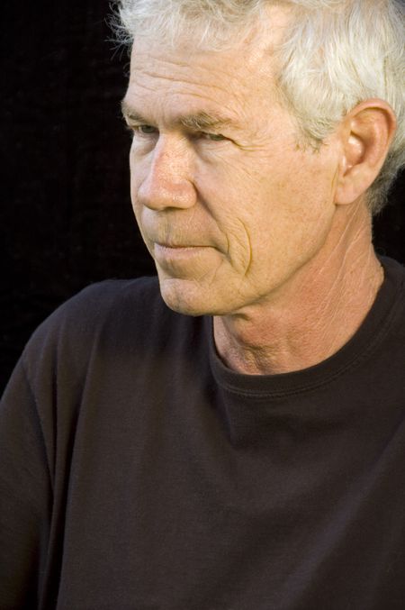 Mature Caucasian man with gray hair, steady gaze, pensive expression, dark background