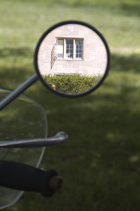 Rearview mirror on motorcycle in shade reflects window of university building in sunlight
