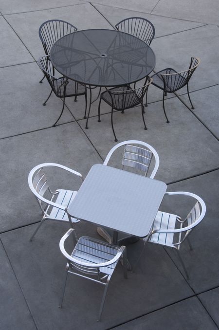 Two sets of metallic cafe tables and chairs, one silver one black, on gray pavement squares, view from above